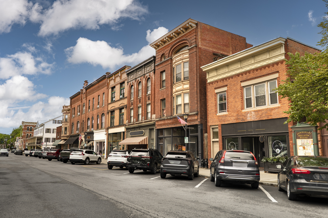 A historic American downtown street lined with brick storefronts, awnings, and parked cars under a partly cloudy sky